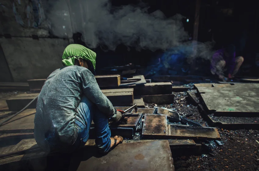 A worker sits among metal parts with smoke rising in a dockyard workshop.