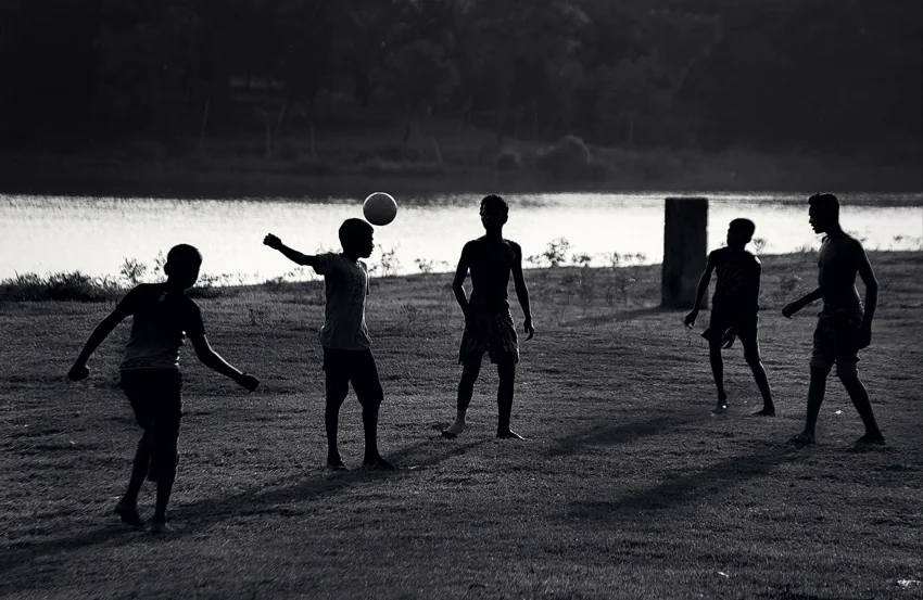 Children play football in silhouette beside water.